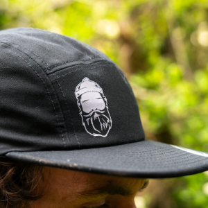 Man wearing 5 panel black cap with bearded men adventures logo.