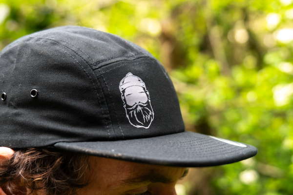 Man wearing 5 panel black cap with bearded men adventures logo.