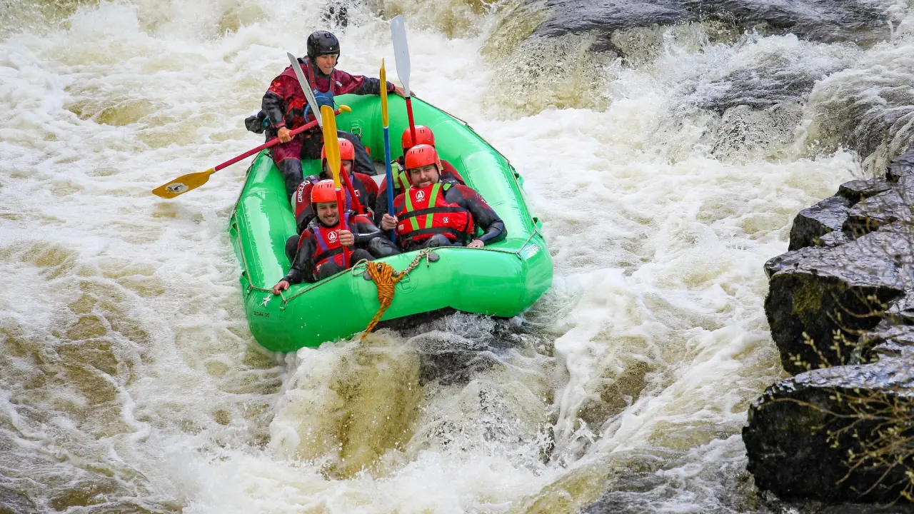 A raft of six paddlers in red helmets punching through a rapid on the River Dee